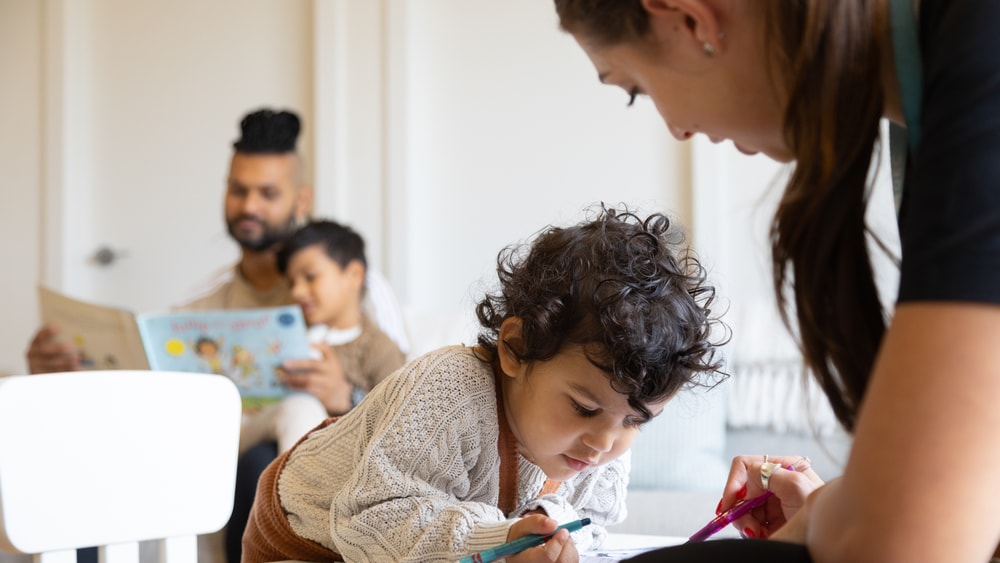 QEC clinical on home visit drawing with child, parent in background.