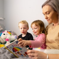 Parent reading book with two children on bed at home.