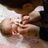 Baby laying on back with dummy in mouth, parent leaning over and looking at baby.