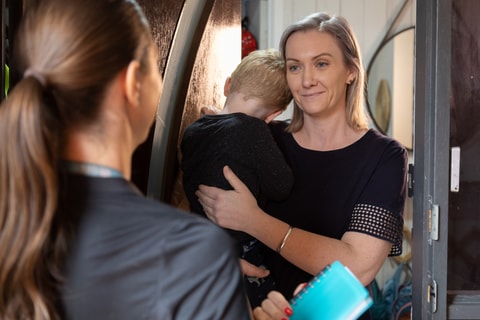 QEC clinician on home visit at front door, parent on other side of door holding child and smiling.