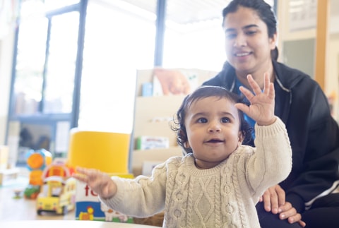 Baby raising hands, parent sitting behind and smiling, in QEC PlayRoom.