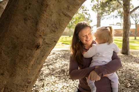 Mum holding toddler up near tree and smiling.